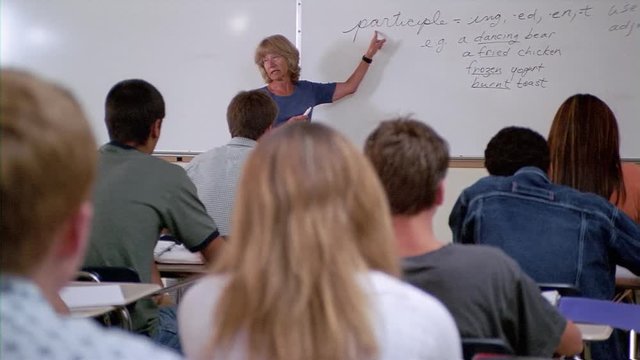 Teacher at whiteboard explaining grammar terms, camera pans left from rear of room