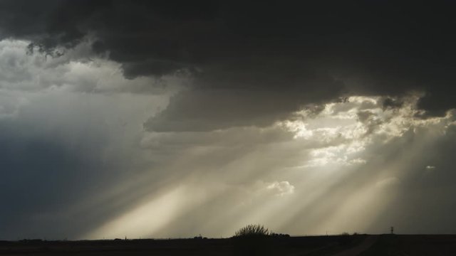 Time-lapse dark clouds pierced by sunbeams glide across the prairie at dusk