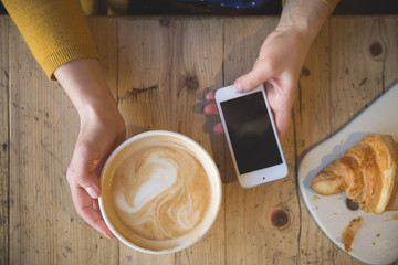 Above view of female hands holding hot cup of coffee  and with smart phone with on wooden table. Coffee break at work. Cappuccino while working.