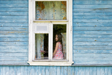 Sad bored little girl sitting on the sill and looking out the country house window. Outside view