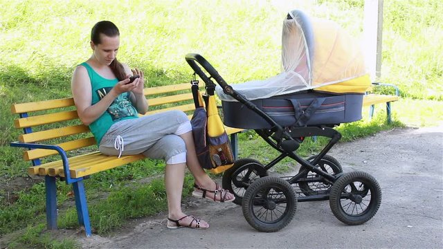 young mother sitting in the park rolls the stroller and looking at smartphone