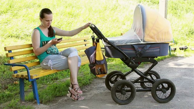 young mother sitting in the park rolls the stroller and looking at smartphone