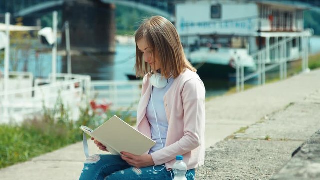 Young adult woman with headphones reading book outdoors smiling at camera