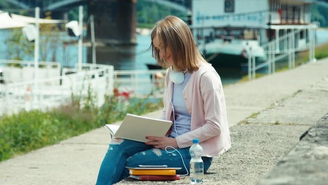 Young adult woman with headphones reading book outdoors near river. Smiling at camera