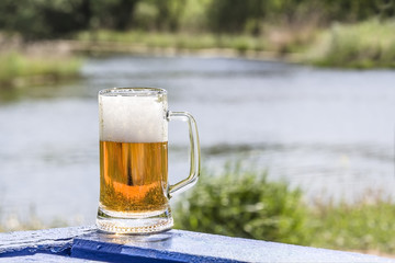 Mug of frothy beer on the table near lake