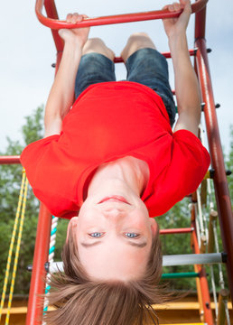 Low Angle View Of Cute Teen Boy Wearing Red Tshirt Hanging Upside Down From A Climbing Frame In A Playground Looking At Camera Smiling