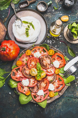 Caprese tomatoes mozzarella salad in aged metal plate with homemade chees and ingredients on dark rustic background, top view