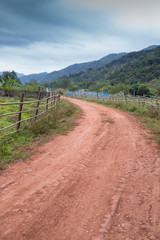 Fototapeta na wymiar red dirt rough pathway road along bamboo fence lead to farm with mountain view, green environment and sky