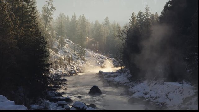 Wintry river flowing from foreground shade toward a sunny bend below a rustic lodge