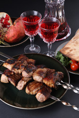 Healthy barbecued lean cubed pork kebabs served with tomato,pita, greens, garnet and two glasses of red drink, close up view on a dark background