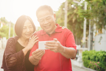 Happy senior Asian couple laughing tigether with mobile smartphone. Warm tone photo with sunlight