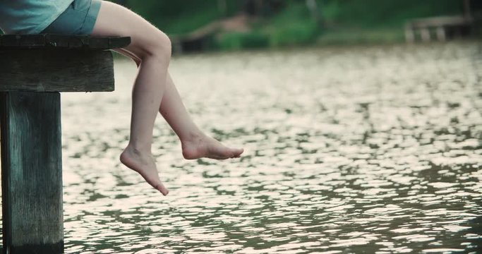 Woman relaxes by the lake sitting on the edge of a wooden jetty , swing one's feet near the water surface. Sunny joyful summer day or evening concept.