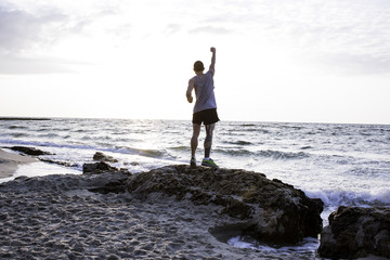Sporty man stand on the rock with rised in air fist sunrise on the beach