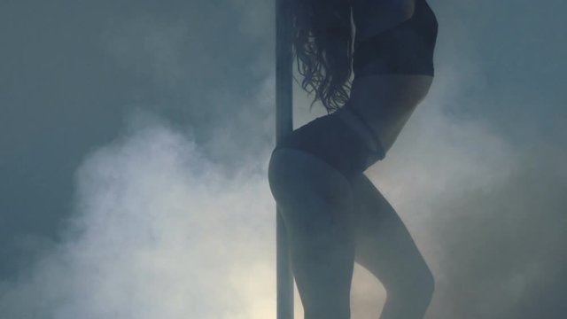 Young slim sexy blonde pole dance woman exercising in studio. Smoke effect on background. Attractive sexy woman pole dancer performing against grey background. Slow motion. 