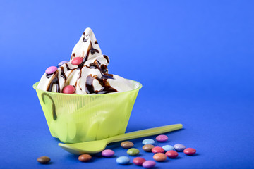 frozen yogurt with chocolate and chocolate candy topping on blue background with green spoon and chocolate candy