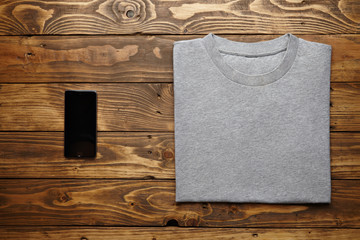 Blank grey t-shirt accurately folded near black smartphone gadget on rustic wooden table top view