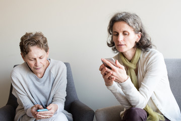 Older mother and middle aged daughter seated next to each other concentrating on smart phones
