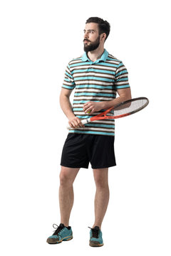 Young adult bearded tennis player holding ball and racket looking away. Full body length portrait isolated over white studio background.