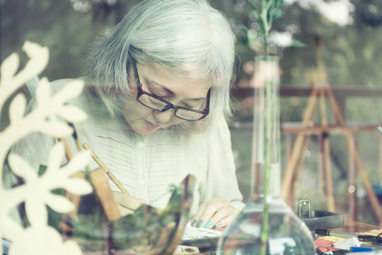 Asian Senior Woman Painting At Her Home Studio, View Through Win