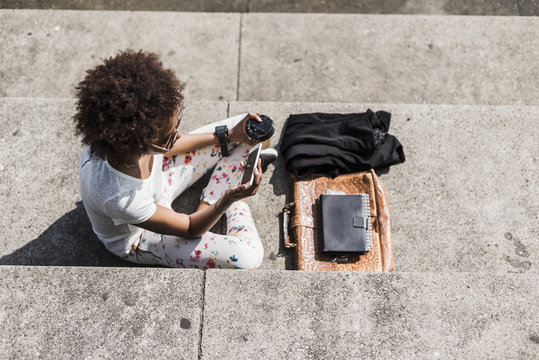 Young woman with coffee to go sitting on stairs looking at her smartphone