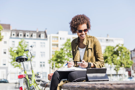 Smiling young woman sitting on a bench using smartphone and digital tablet