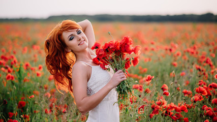 Naklejka premium beautiful young red-haired woman in poppy field holding a bouquet of poppies
