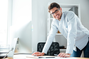 Cheerful young businessman standing and using computer in office