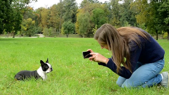 young attractive happy woman sits on the ground and takes pictures of french bulldog in the park