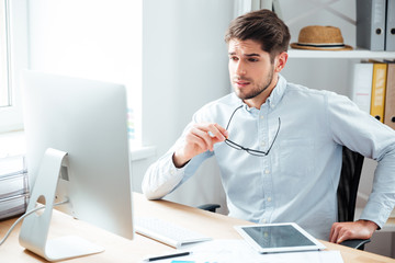 Fototapeta premium Handsome businessman holding eyeglasses and looking at computer monitor