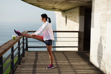 Asian sporty woman stretching legs for warming up before running or exercising outdoor. Female athlete on fitness training towards the sea.