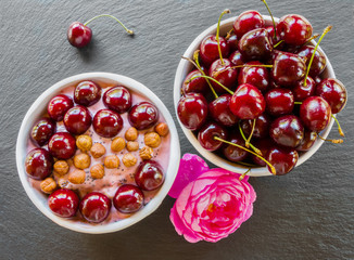 Breakfast bowl with yogurt,  granola or muesli or oat flakes, fresh cherries and nuts. Black stone background, pink rose flower. Top view.