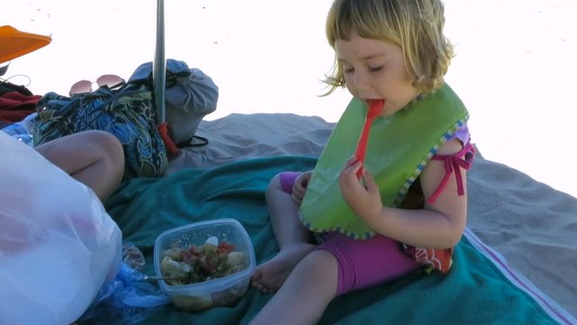 little baby next to mother sitting down of parasol at beach eating tuna and green bean salad from tupperware with orange fork
