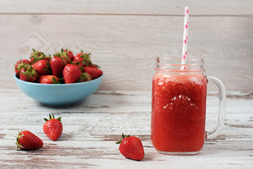 Fresh juice, shake, milkshake of strawberries in a mason jar with a straw. Pile of juicy ripe organic fresh strawberries in a large blue bowl. Light rustic wooden background