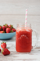 Fresh juice, shake, milkshake of strawberries in a mason jar with a straw. Pile of juicy ripe organic fresh strawberries in a large blue bowl. Light rustic wooden background