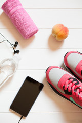 Red sneakers with towel, peach, bottle with water, smartphone with headphones on white wooden background indoors. Sports and fitness background, healthy lifestyle concept.