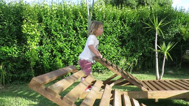 slow motion two years age blonde cute baby with white shirt jumping and playing on wooden hammock in green garden plants

