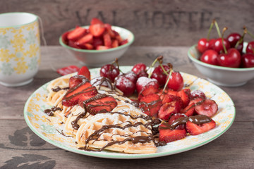 Soft Belgian heart shaped waffles with cherries and strawberries, chocolate topping and powdered sugar on yellow plate. Wooden background.
