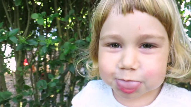 slow motion of close up two years age blonde baby girl face talking, looking, teasing, sticking out tongue and hitting camera in green garden plants background
