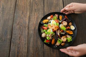 Hands holding plate with seafood mix salad on wooden background. Taking from wooden table plate with seafood salad hands. free space for commercials