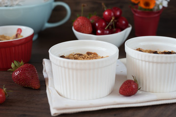 Cherry, strawberry crumble dessert in white bowls. Cherries, strawberries, flowers behind. Dark wood background