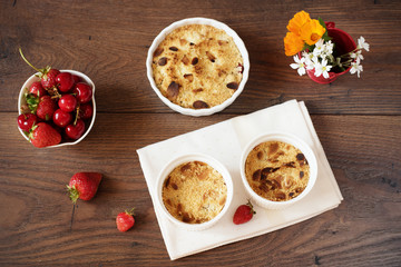 Cherry, strawberry crumble dessert in white bowls. Cherries, strawberries, flowers behind. Dark wood background, top view