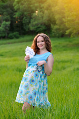 Fototapeta na wymiar Happy pregnant girl standing in a meadow at sunset, smiling and holding baby shoes in her hand. Vertical orientation