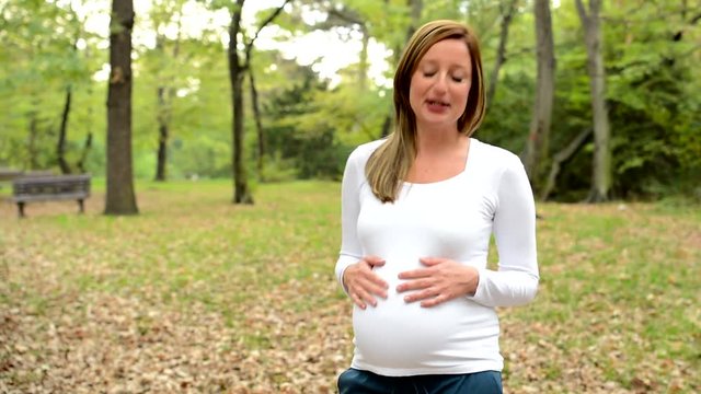 Young pretty pregnant woman talks to camera about something in park