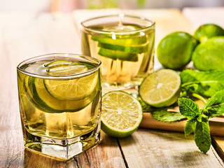 Country drink. On wooden boards are two glasses with green transparent drink and half lime glue bowl. A drink number twenty mojito with mint leaf on sunlight . Country life. Light background.