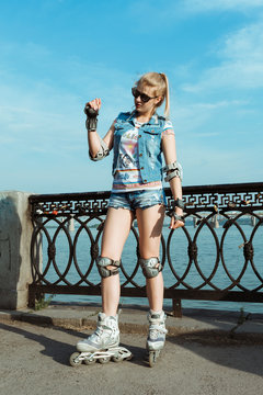 Young fit women girl in sunglasses, jeans shorts and a jacket on roller skates riding outdoors on river embankment, on the promenade