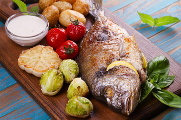 Broiled dorado fish with brussels sprouts, tomatoes, garlic, young potato and greens on cutting board