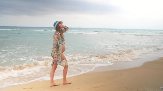 Stylish pregnant woman in swimsuite and tunic walking on a sandy beach along sea waves.