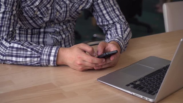 Male typing fast on mobile. Guy holding phone above wooden desk and chatting in social net. Model wearing in casual plaid shirt. Unrecognisable person details.