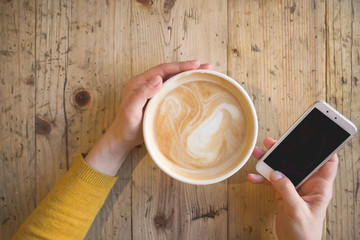 Above view of female hands holding hot cup of coffee  and with smart phone with on wooden table. Coffee break at work. Cappuccino while working.