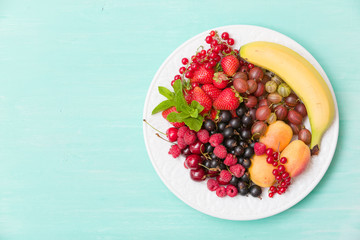 Assortment of juicy fruits on white plate and turquoise table background. Banana, strawberries, blueberries, raspberries, gooseberry, blackcurrant decorated with mint for summer dessert or snack.